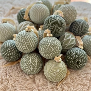 Pu&ograve; includere: Una collezione di addobbi natalizi verdi con vari motivi in rilievo, tra cui forme geometriche, foglie e volute. Gli addobbi sono disposti in un mucchio su una superficie soffice di colore beige.