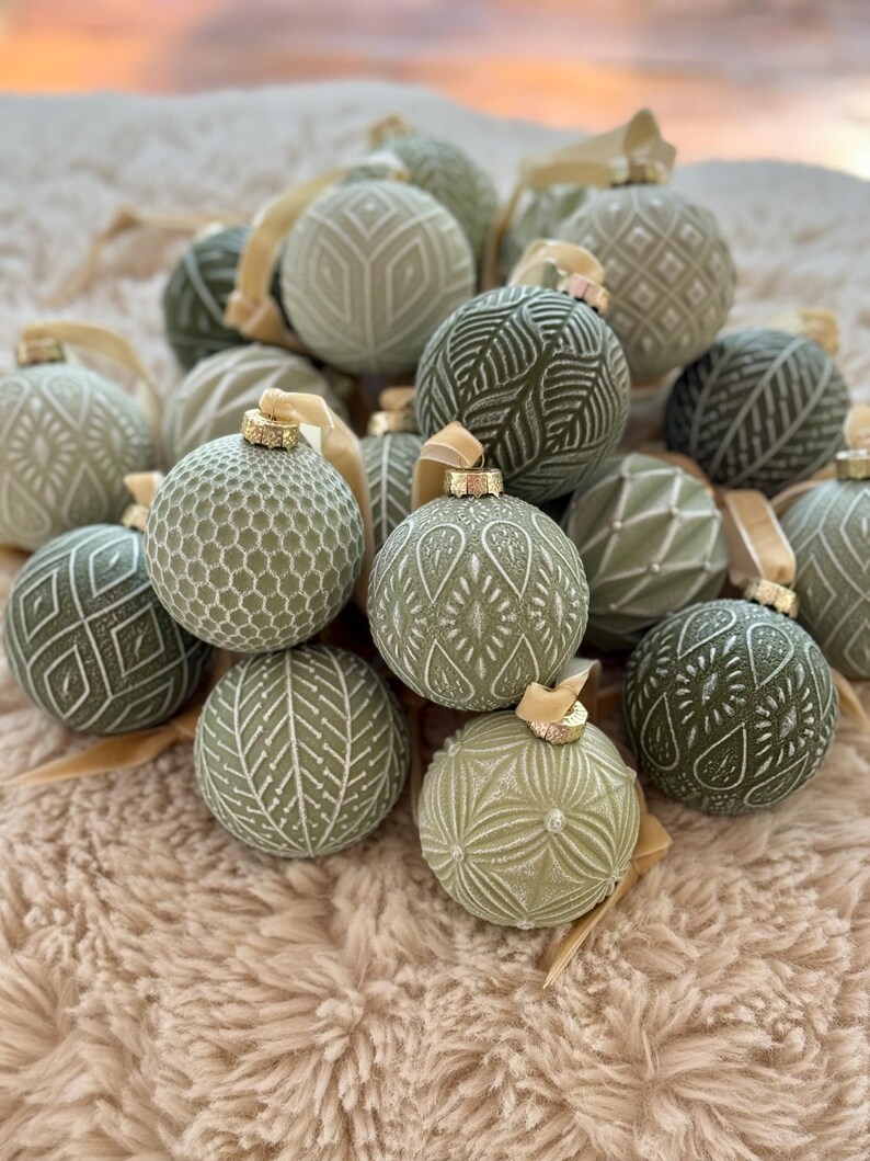 Pu&ograve; includere: Una collezione di addobbi natalizi verdi con vari motivi in rilievo, tra cui forme geometriche, foglie e volute. Gli addobbi sono disposti in un mucchio su una superficie soffice di colore beige.