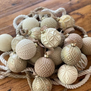 Pu&ograve; includere: Una collezione di addobbi natalizi color crema e oro con intricati disegni in rilievo. Gli addobbi sono disposti su una ghirlanda di corda e una superficie in legno.