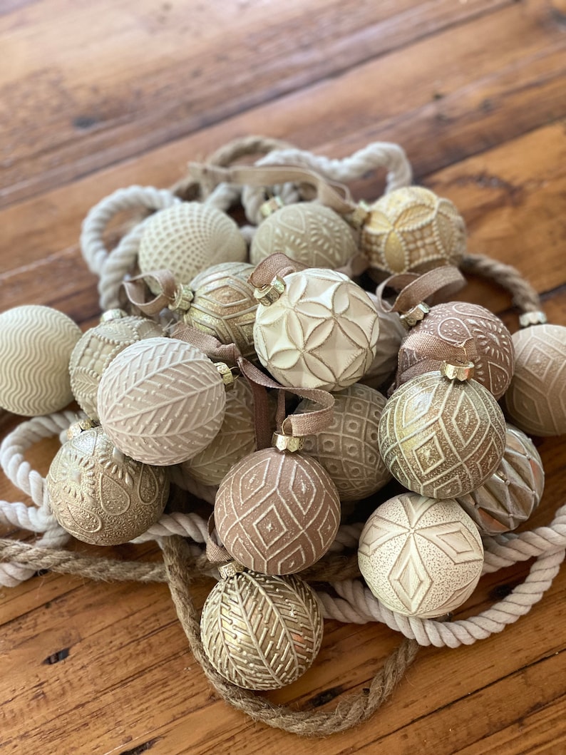 Pu&ograve; includere: Una collezione di addobbi natalizi color crema e oro con intricati disegni in rilievo. Gli addobbi sono disposti su una ghirlanda di corda e una superficie in legno.