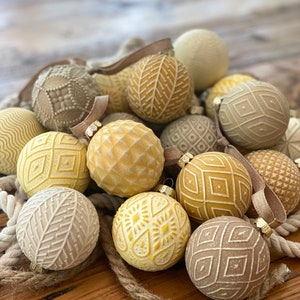 Pu&ograve; includere: Una collezione di 15 palline di Natale rotonde, crema, beige e gialle con vari motivi in rilievo. Le palline sono disposte in un mucchio su una superficie di legno.