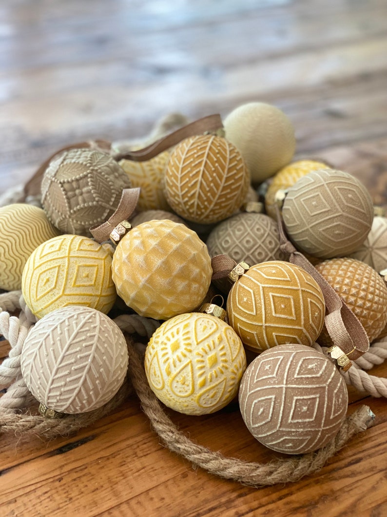 Pu&ograve; includere: Una collezione di 15 palline di Natale rotonde, crema, beige e gialle con vari motivi in rilievo. Le palline sono disposte in un mucchio su una superficie di legno.