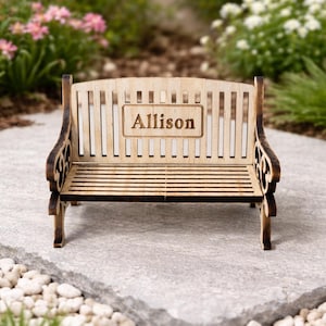 May include: A small wooden bench with the name "Allison" engraved on the backrest. The bench features a slatted seat and back, with decorative curved armrests. The wood has a natural finish with darker edges. The bench is outdoors.