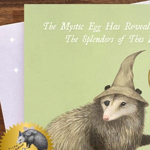 Może przedstawiać: Kapryśna kartka z życzeniami z opossem w kapeluszu czarodzieja i przepaską na oku. Na karcie widnieje napis "The Mystic Egg Has Revealed The Splendors of This". Złota pieczęć z kresk&oacute;wkowym opossem znajduje się w lewym dolnym rogu.