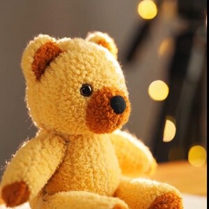 Peut inclure: Un ours en peluche avec un corps jaune clair et des accents bruns sur les oreilles, les pattes et le museau. L'ours a des yeux noirs en forme de boutons et un nez noir cousu. Le jouet est assis.