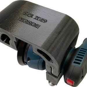 May include: A black 3D printed sander attachment for a Bosch GEX 125 sander. The attachment is designed to hold the sander in a specific position.