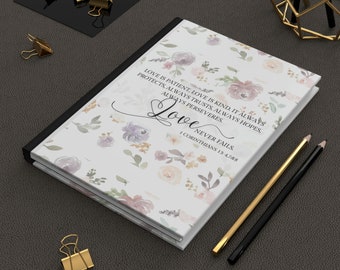 Puede incluir: Un diario con estampado floral con la palabra "Love" y el versículo bíblico 1 Corintios 13:4-8 impresos en la portada. La portada presenta un diseño de flores en colores pastel. También se ven lápices y clips dorados y negros.