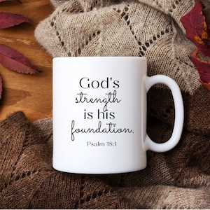 Może przedstawiać: Biały ceramiczny kubek z napisem "God's strength is his foundation. Psalm 18:1" czarną czcionką. Kubek ma biały uchwyt i jest umieszczony na tle brązowego, dzierganego koca i jesiennych liści.