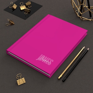 May include: A bright pink journal with the name "Jasmine Jones" printed on the cover. Gold and black pencils and gold binder clips are arranged around the journal on a dark gray surface.