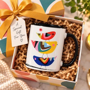 Puede incluir: Una taza de cer&aacute;mica blanca con asa y borde negros, que presenta un gr&aacute;fico colorido de tres p&aacute;jaros apilados en rojo, amarillo y azul. La taza est&aacute; en una caja de regalo con relleno de papel marr&oacute;n y una etiqueta que dice A Special Gift For You.