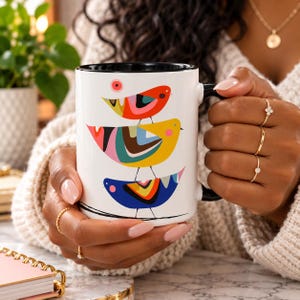 Puede incluir: Una taza de cer&aacute;mica blanca con interior y asa negros, que presenta una ilustraci&oacute;n gr&aacute;fica de tres p&aacute;jaros apilados en colores vibrantes rojo, amarillo y azul con patrones abstractos, sostenida por manos adornadas con varios anillos de oro.