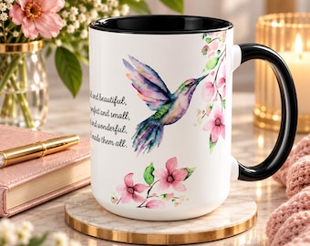 Puede incluir: Una taza de cerámica blanca con asa y borde interior negros, que presenta una ilustración en acuarela de un colibrí en vuelo, detalles florales rosas y el texto en negro: "...and beautiful, ...perfect and small, ...and wonderful, ...made them all."