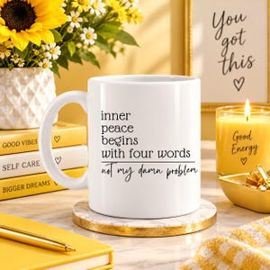 Może przedstawiać: Biały ceramiczny kubek z czarnym napisem: inner peace begins with four words, poniżej którego znajduje się pozioma linia oraz fraza: not my damn problem. Kubek posiada zakrzywione ucho i stoi na okrągłej podstawce z marmurowym wzorem.