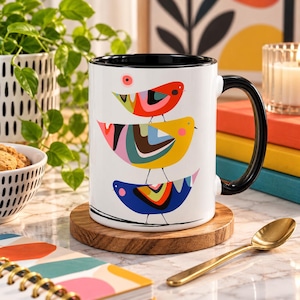 Puede incluir: Una taza de cer&aacute;mica blanca con interior y asa negros, que presenta una pila vertical de tres p&aacute;jaros estilizados en rojo, amarillo y azul con coloridos patrones geom&eacute;tricos en sus cuerpos, apoyada sobre un posavasos de madera redondo.