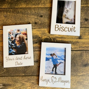 May include: Three white wooden picture frames with photos inside. One frame has the name "Biscuit" engraved, another has "Your text here Date", and the third has "Humza ♡ Maryam". The frames are on a wooden surface.