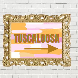 Op de afbeelding: Een gouden lijst met een abstracte achtergrond in roze en oranje. Het bord zegt "TUSCALOOSA" in bruine letters met een turquoise omtrek en een gouden pijl die naar rechts wijst.