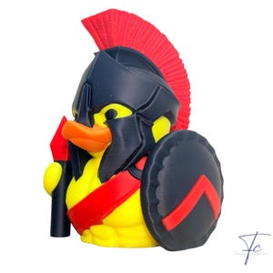 May include: A yellow rubber ducky wearing a black Spartan helmet with a red plume, a red chest strap, and a black shield with a red chevron. The ducky holds a black spear with a red tip. The ducky has an orange beak.