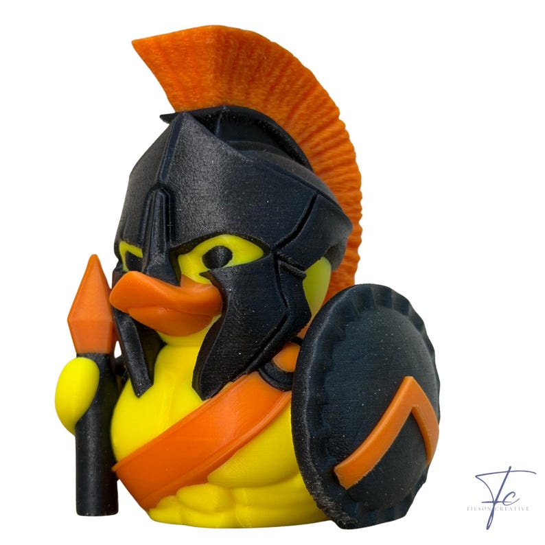 May include: A yellow duck figurine styled as a Spartan warrior, featuring a black helmet with an orange crest, shield, and spear. The duck has an orange beak and accents, creating a unique and playful design.