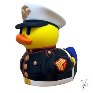 May include: A yellow rubber duck wearing a U.S. Marine Corps uniform, including a white hat with gold emblem, black jacket with gold buttons, and a red and gold patch. The duck has an orange beak.
