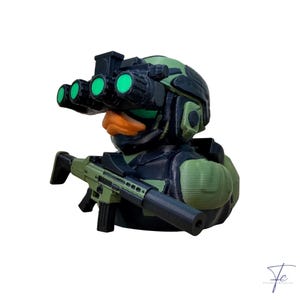 Könnte beinhalten: Eine taktische Gummi-Ente-Figur, die einen grünen Kampfhelm mit einer schwarzen Nachtsichtbrille mit vier leuchtend grünen Linsen trägt. Die Ente hat einen orangefarbenen Schnabel und ist mit einem schwarz-grünen Sturmgewehr mit Schalldämpfer ausgestattet.
