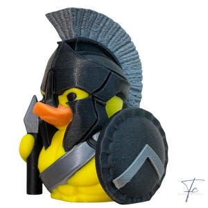 May include: A yellow rubber ducky in a black Spartan helmet, shield, and spear. The duck has an orange beak. The shield has a silver chevron design. The background is white.
