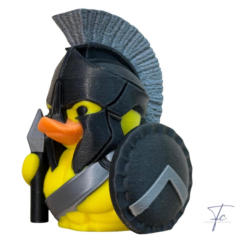 May include: A yellow rubber ducky in a black Spartan helmet, shield, and spear. The duck has an orange beak. The shield has a silver chevron design. The background is white.
