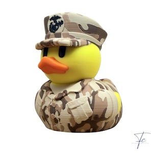 May include: A yellow rubber duck figurine wearing a desert camouflage military uniform and a matching peaked cap. The cap features a dark brown emblem of an eagle, globe, and anchor. The duck has a bright orange beak and black eyes.