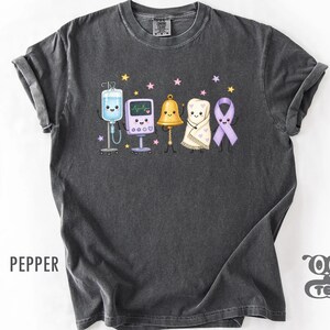 May include: A charcoal gray short-sleeve t-shirt featuring a graphic of five cute, smiling characters: an IV bag, a heart monitor, a gold bell, a blanket, and a purple awareness ribbon, all surrounded by small pink and yellow stars.
