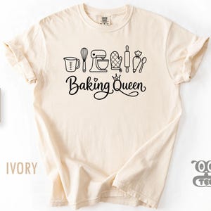 May include: An ivory short-sleeve t-shirt featuring a black graphic print of various baking tools including a measuring cup, whisk, stand mixer, oven mitt, rolling pin, piping bag, and spoon, with the text "Baking Queen" written below.