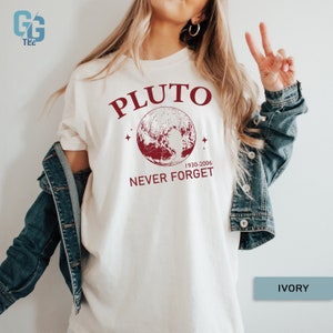 May include: A white t-shirt with a red graphic that says "Pluto 1930-2006 Never Forget" with a red illustration of Pluto. The text "IVORY" is in the bottom right corner of the image.