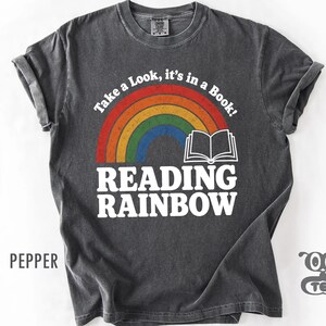 May include: A charcoal gray short-sleeve t-shirt featuring a distressed rainbow graphic above an open book icon. The text reads Take a Look, it's in a Book! and READING RAINBOW in white capital letters. The shirt has a crew neckline and a vintage wash.
