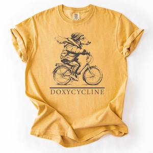 May include: A mustard yellow t-shirt with a graphic of a cartoon animal wearing a winter coat and riding a bicycle. The text "DOXYCYCLINE" is printed below the graphic.