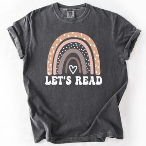 May include: A dark gray t-shirt with a rainbow design and the text "Let's Read" in white. The rainbow is made up of pink, brown, and gray stripes with white polka dots.