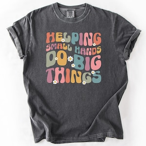 May include: A dark gray t-shirt with the text "Helping Small Hands Do Big Things" in a retro font with colorful letters and white flowers.