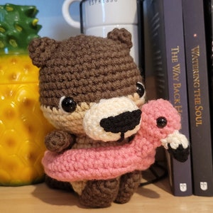 May include: A brown crocheted bear holding a pink inflatable flamingo float. The bear has black eyes and a black nose. The bear is sitting on a wooden surface.