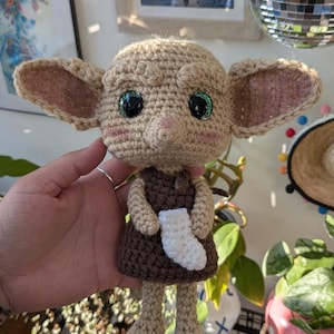 May include: A tan crochet doll with large green eyes, prominent ears, and pink cheeks, wearing a dark brown dress and holding a small white sock. The doll features textured yarn stitching, a rounded head, and slender limbs in a handcrafted style.