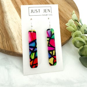 May include: A pair of rectangular earrings with a colorful stained glass design. The earrings feature a vibrant mix of red, pink, blue, green, and yellow hues, outlined in black. The earrings are displayed on a white card with the text "JUST JEN - Handcrafted Jewelry".