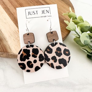 May include: A pair of wooden earrings with a leopard print design and a brown square accent. The earrings are handmade and feature silver hooks.