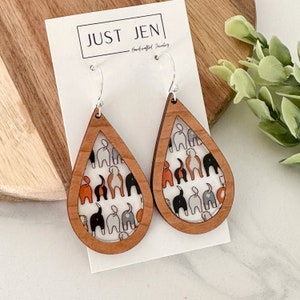 May include: A pair of wooden teardrop-shaped earrings with a white background and a pattern of dogs' rear ends in black, gray, and orange. The earrings are hanging from silver hooks and are labeled "JUST JEN Handcrafted Jewelry".