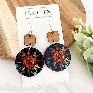 May include: A pair of wooden earrings with a sun design. The earrings are made of wood and have a silver hook. The sun design is in shades of orange, yellow, and blue. The earrings are on a white card with the text "JUST JEN - Handcrafted Jewelry".