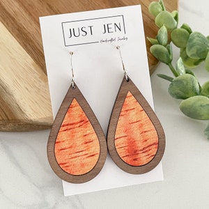 May include: A pair of teardrop-shaped wooden earrings with a walnut wood frame and a bright orange wood inlay. The earrings are hanging from silver hooks. The earrings are on a white card with the text "JUST JEN - Handcrafted Jewelry".