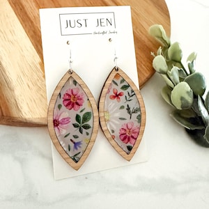 May include: Wooden teardrop earrings with a floral design. The earrings feature a clear center with pink and red flowers, green leaves, and small blue flowers. The earrings are framed with light-colored wood and have silver hooks. The text "JUST JEN" is visible on the card.