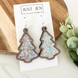 May include: A pair of silver tone dangle earrings featuring a wood cut-out of a Christmas tree filled with iridescent glitter stars. The earrings are on a white card with the text "JUST JEN - Handcrafted Jewelry".