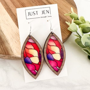 May include: A pair of wooden earrings with a stained glass design in shades of red, pink, yellow, and purple. The earrings are shaped like teardrops and have silver hooks.