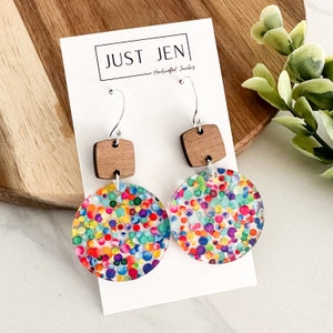 May include: A pair of colorful confetti earrings with a wood accent. The earrings are made of clear resin with a confetti pattern in shades of red, orange, yellow, green, blue, purple, and pink. The wood accent is a small square piece of wood with a natural finish.