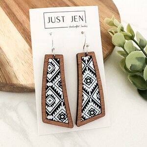 May include: A pair of wooden earrings with a black and white geometric pattern. The earrings are made of wood and have silver hooks. The earrings are called "JUST JEN - Handcrafted Jewelry".