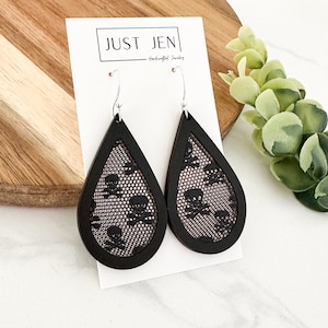 May include: A pair of black teardrop-shaped earrings with a lace pattern and skull design. The earrings are made of wood and have silver hooks.