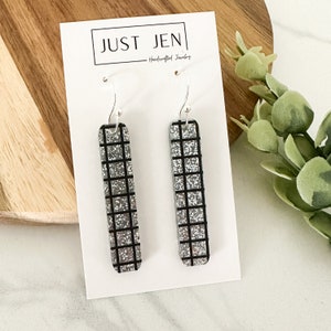 May include: Silver glitter earrings with a black grid pattern. The earrings are on a white card with the text "JUST JEN - Handcrafted Jewelry".