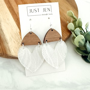 May include: A pair of earrings with a wooden half-circle top and a heart-shaped bottom. The heart-shaped portion is white with a subtle, repeating leaf pattern. The earrings are displayed on a white card with the text "JUST JEN - Handcrafted Jewelry".