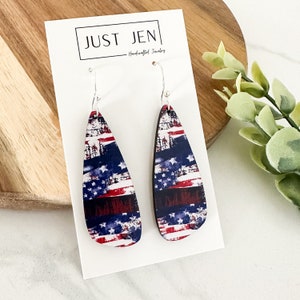 May include: A pair of teardrop-shaped earrings with a distressed American flag design. The earrings are made of wood and have silver hooks.